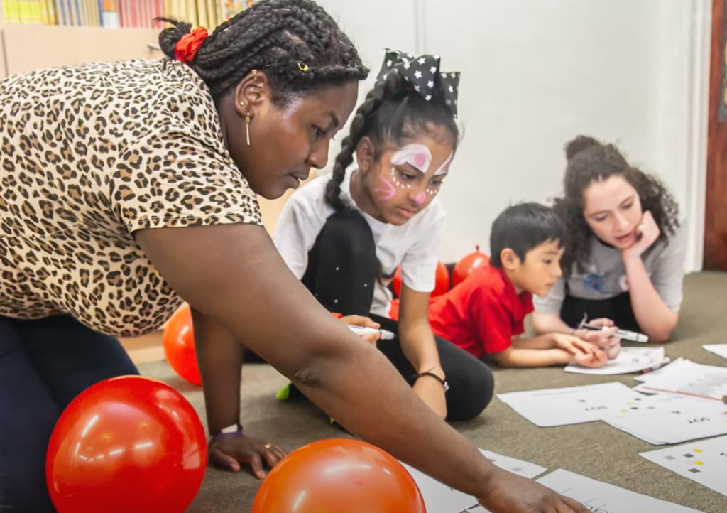 A female creative coach points to something on a page while a young female student with face paint watches. They are surrounded by red balloons.