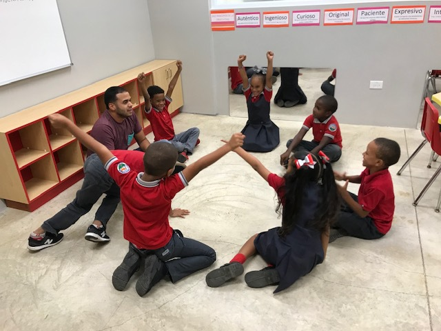 Six small children sit in a circle with their Creative coach. Some of the students have their hands in the air with excitement.