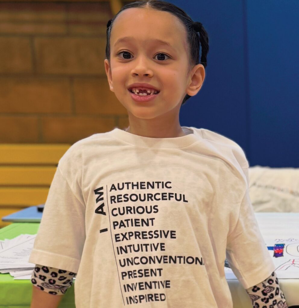 A smiling student wearing a T-shirt listing the 10 Principles of Creativity: authentic, resourceful, curious, patient, expressive, intuitive, unconventional, present, inventive, inspired